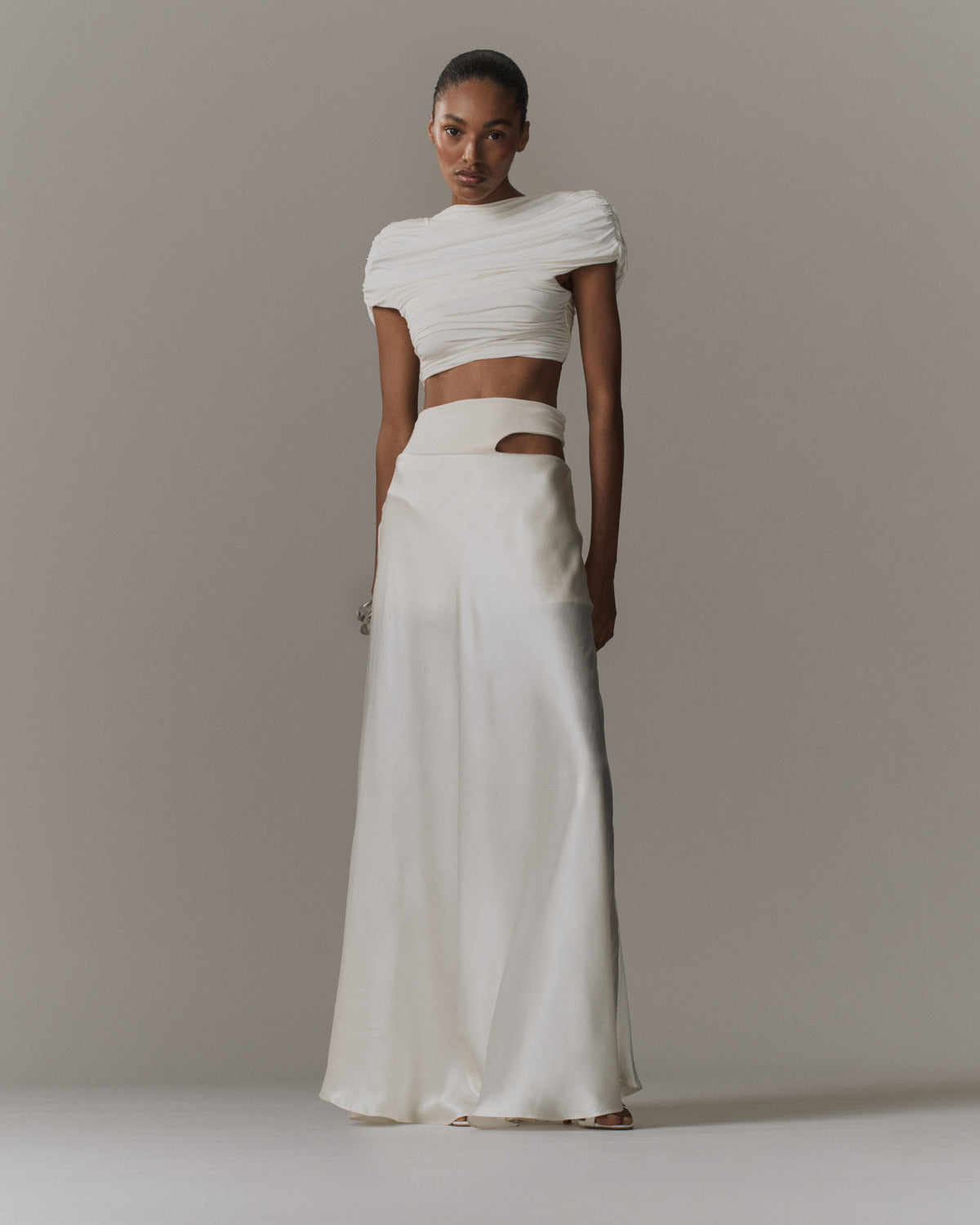 A model wearing a two-piece white outfit consisting of a ruched short-sleeve crop top and a high-waisted, wide-leg satin skirt, set against a plain light gray studio background.