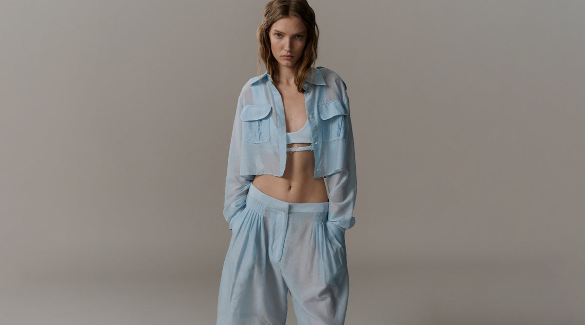 A model wearing a light blue cropped button-up shirt and matching wide-leg pants over a matching light blue bralette, set against a plain gray studio background.