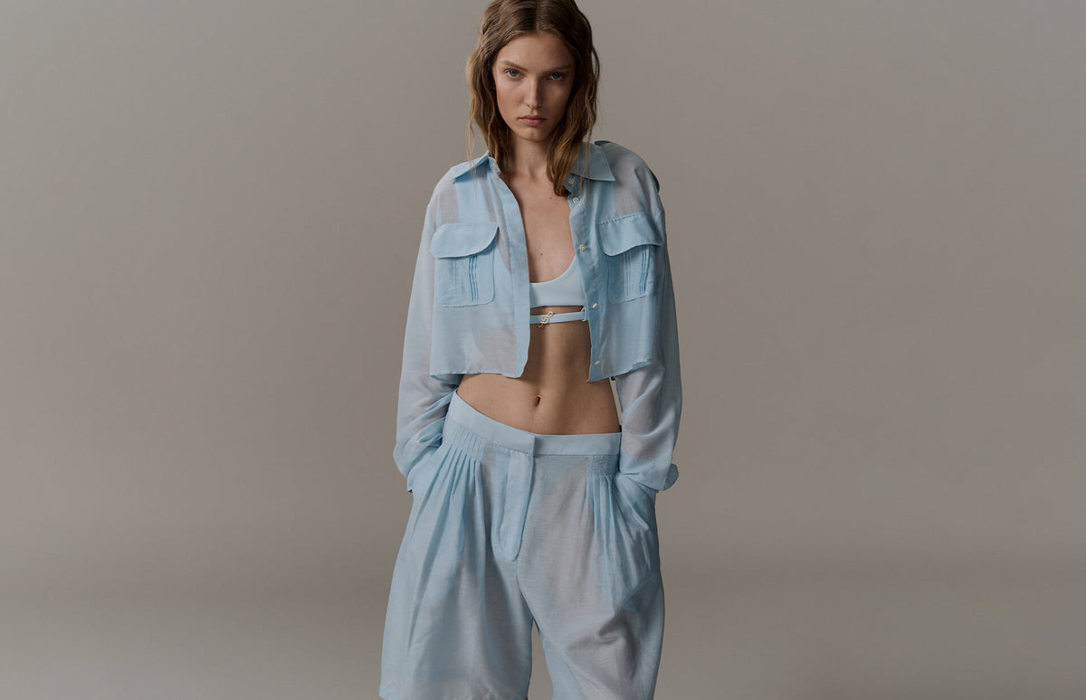 A model wearing a light blue cropped button-up shirt and matching wide-leg pants over a white bralette, standing against a neutral gray studio background.